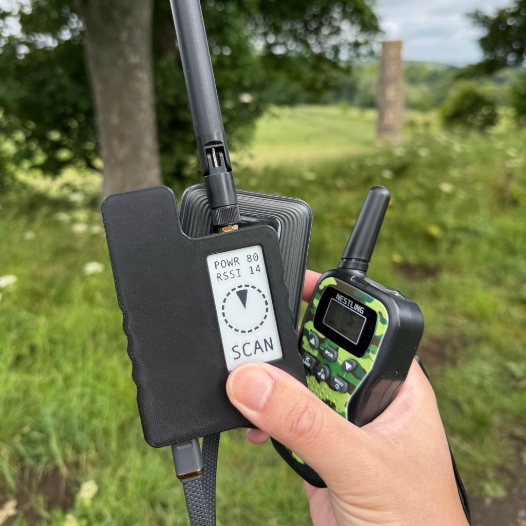 Adrian's hand, holding version 2 of the MFD, a battery pack and a walkie talkie. The battery pack is connected to the MFD. Behind Adrian's hand is the beautiful green scenery of Cammo Estate and, out of focus, Cammo Tower.