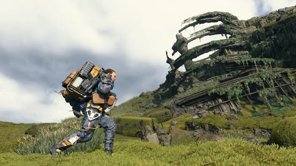 Screenshot from Death Stranding, with a man in a high tech exoskeleton carrying big packages on his back, striking through a post-apocalyptic landscape