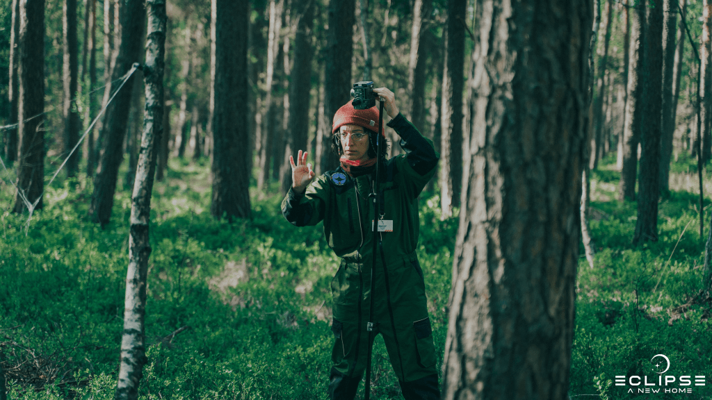 A person in a sci-fi jumpsuit in a forest setting up a device on a tripod, signalling OK to another person