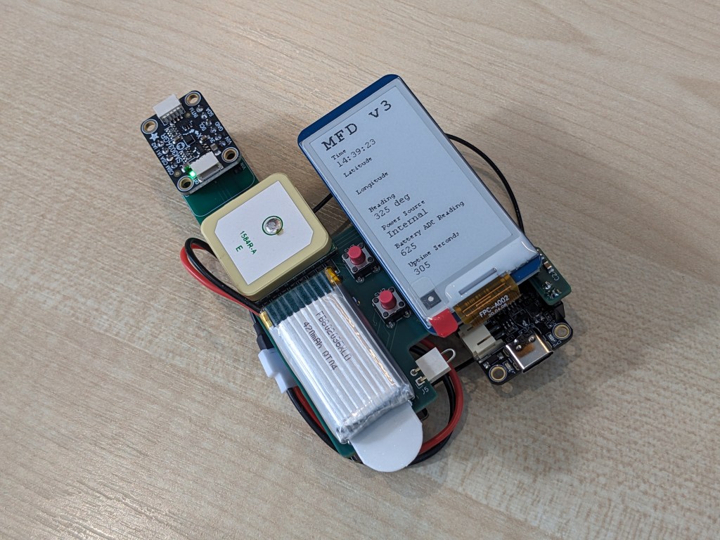 An electronic device without an enclosure. The device has an ePaper display which shows some text for verifying the correct operation of the device. The title on the screen is "MFD v3". The screen also shows the current time, a compass heading, an uptime in seconds and a "battery ADC reading".